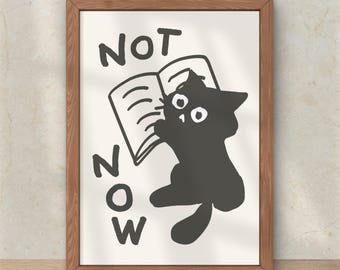 Puede incluir: Un cuadro enmarcado en madera con una ilustración de un gato negro leyendo un libro. El texto "NOT NOW" está escrito en letras negras. El gato está dibujado en un estilo de dibujos animados simple, con ojos grandes.
