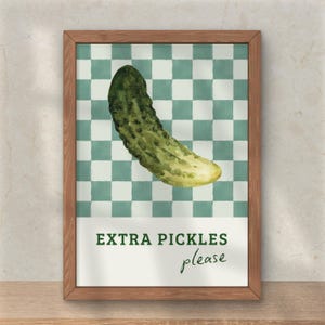 May include: A framed print featuring a watercolor illustration of a green pickle against a checkered background. The text "EXTRA PICKLES please" is displayed below the pickle in a white banner. The frame is made of wood.