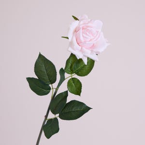 May include: A single artificial rose with a pale pink flower head and green leaves. The rose is on a long, green stem. The background is a plain, light pink colour.