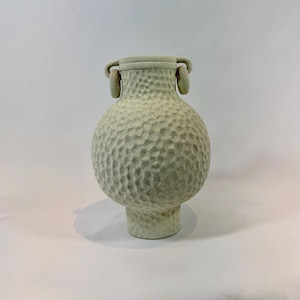Puede incluir: Un jarrón de cerámica verde claro con una superficie texturizada y con hoyuelos. El jarrón tiene un cuerpo bulboso, una base corta de pedestal y dos asas de bucle cerca del borde. Altura aproximada de 25 cm.