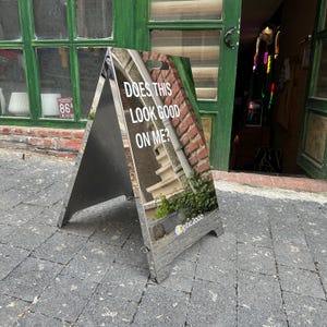 May include: A-frame sign with the text "DOES THIS LOOK GOOD ON ME?" in white. The sign is reflective and shows a mirrored image of the surroundings. The bottom right corner has the "Optic 2000" logo.