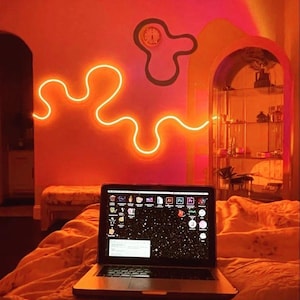 May include: A laptop computer with a bright, colorful desktop background sits on a bed with orange bedding. The room is lit with orange neon lights and a neon light in the shape of a squiggly line is on the wall.