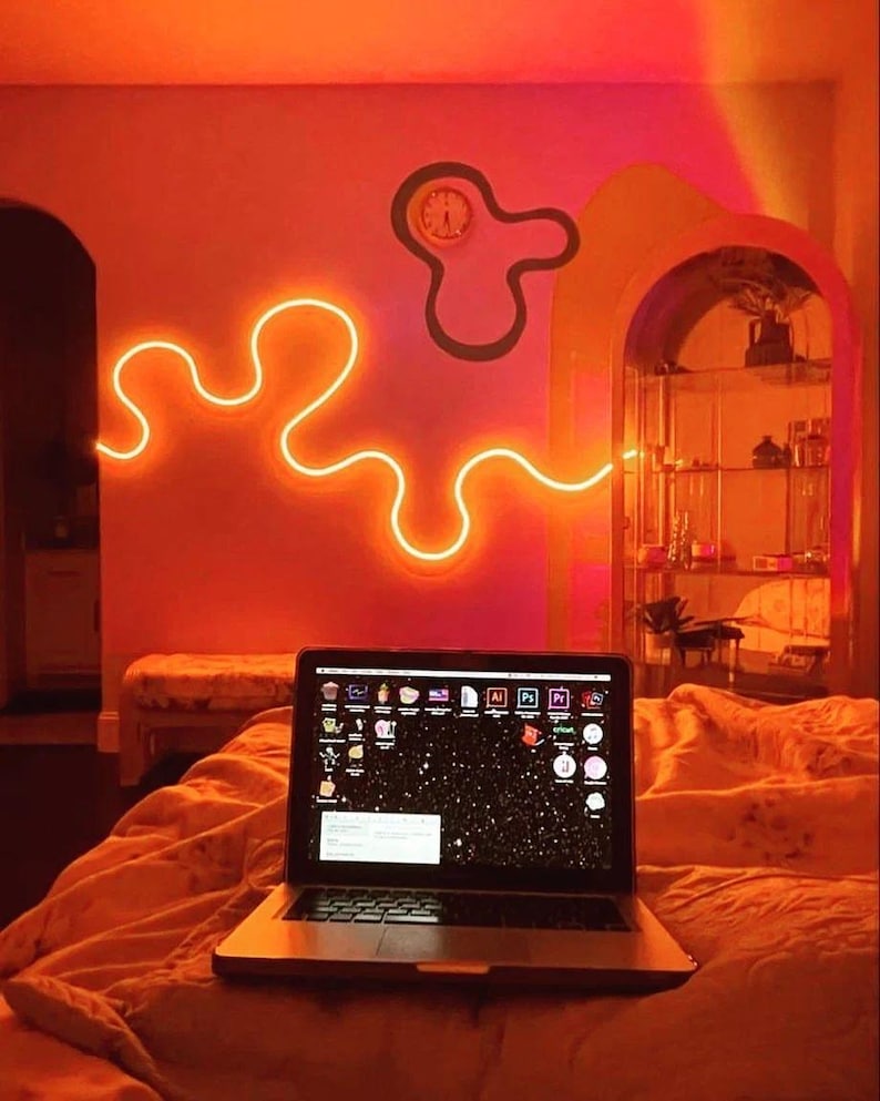 May include: A laptop computer with a bright, colorful desktop background sits on a bed with orange bedding. The room is lit with orange neon lights and a neon light in the shape of a squiggly line is on the wall.