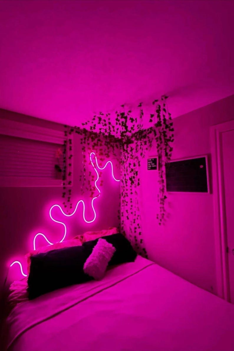 May include: A pink bedroom with a bed, a neon light shaped like a wave, and a wall decorated with hanging greenery.