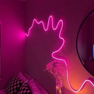May include: A pink neon light shaped like a cloud hangs on a wall in a bedroom. The light is on and illuminating the room. There is a bed with pillows and a nightstand with a candle and a vase of flowers.