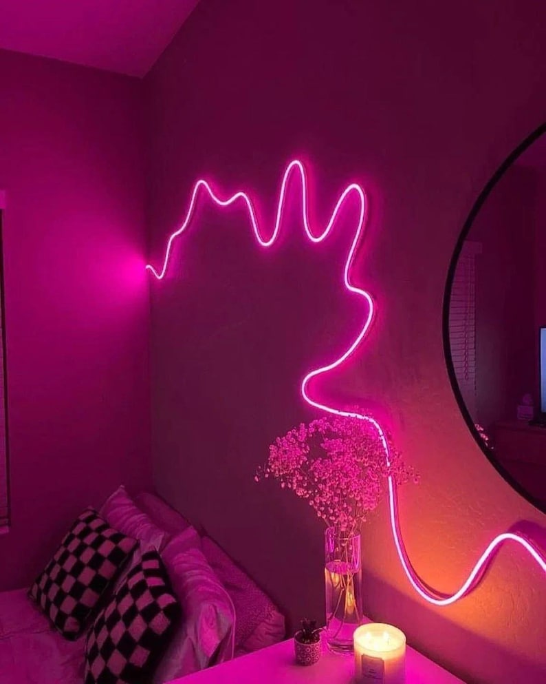 May include: A pink neon light shaped like a cloud hangs on a wall in a bedroom. The light is on and illuminating the room. There is a bed with pillows and a nightstand with a candle and a vase of flowers.