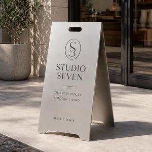 Pode incluir: Uma placa de cavalete prateada com uma alça recortada no topo. A placa apresenta um logótipo circular com as letras SS, o texto STUDIO SEVEN, a frase TIMELESS PIECES MODERN LIVING e a palavra WELCOME impressas a preto na face frontal.