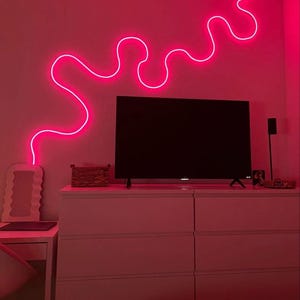 May include: A pink neon light in the shape of a wavy line is mounted on a wall above a white dresser with six drawers. A television is on top of the dresser.