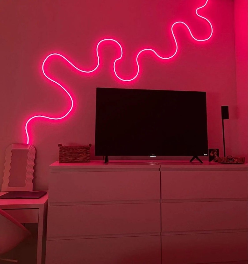May include: A pink neon light in the shape of a wavy line is mounted on a wall above a white dresser with six drawers. A television is on top of the dresser.