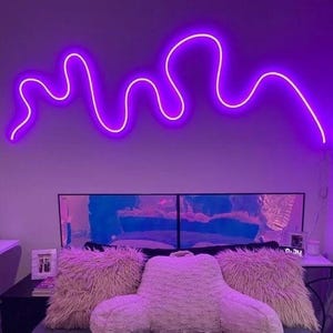 May include: A purple neon light shaped like a wavy line hangs on a wall above a bed with fluffy pillows.
