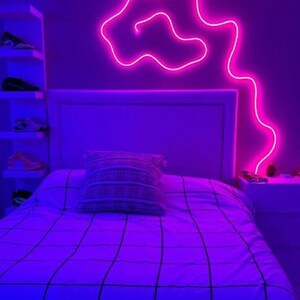 May include: A bedroom with a bed, a white headboard, and a pink neon light shaped like a cloud on the wall. The bed has a white comforter with a grid pattern and a white pillow with a patterned design. The room is lit with purple and blue lights.