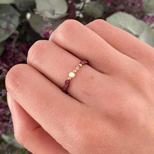 Peut inclure: Une bague de perles avec une perle blanche centrale, flanquée de petites perles roses et bordeaux. Des perles dorées ornent le design. La bague est portée sur un doigt, avec un arrière-plan flou de feuillage violet et vert.