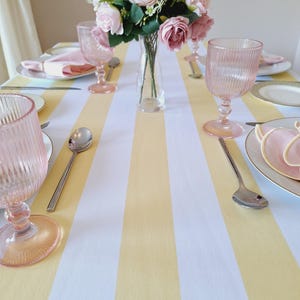 Op de afbeelding: Een tafelsetting met een geel en wit gestreepte tafelloper. Roze waterglazen, borden en servetten omringen een vaas met roze rozen. Zilveren lepels zijn op de loper geplaatst.