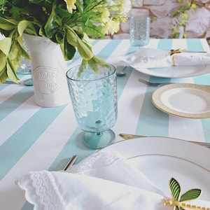 Puede incluir: Una mesa bellamente decorada con un mantel a rayas azul claro y blanco. Platos blancos, servilletas y cubiertos están dispuestos. Un vaso azul claro y un jarrón de flores se suman a la elegante escena. Servilleteros con forma de libélula.