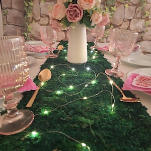 May include: A green moss table runner with a string of fairy lights, two gold spoons, and pink glasses. A bouquet of pink roses in a white vase is in the centre of the table runner.