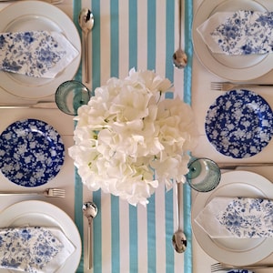 Puede incluir: Una mesa puesta con platos blancos, platos laterales azules con motivos florales y servilletas a juego. Cubiertos plateados y vasos teñidos de azul rodean un centro de mesa de flores blancas. Un camino de mesa a rayas azules y blancas.