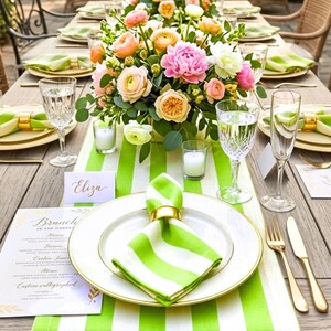 May include: A dining table setting featuring a green and white striped table runner, white plates with gold rims, green and white striped napkins held by gold rings, gold cutlery, crystal glassware, and a centerpiece of pink, peach, and white flowers.