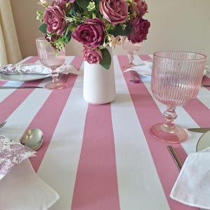 May include: A dining table setting with a pink and white striped tablecloth. A bouquet of burgundy and pink roses sits in a white vase. Pink glassware, silverware, and napkins complete the elegant table arrangement.