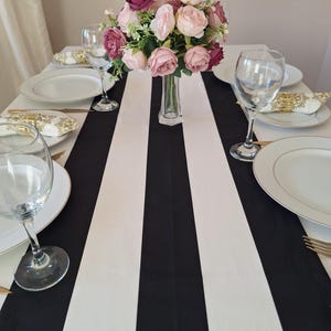 May include: A black and white striped tablecloth with a vase of pink and burgundy flowers in the center. There are four place settings with white plates, gold silverware, and clear wine glasses.