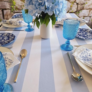 May include: A table setting with a blue and white striped tablecloth, featuring blue goblets, floral plates, and silver spoons. A white vase holds a bouquet of light blue hydrangeas, creating an elegant and coordinated look.