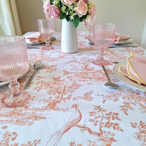 Puede incluir: Una mesa puesta con un mantel blanco con un estampado de toile de Jouy naranja. Copas, servilletas y un jarrón con flores rosas componen un ambiente elegante. También se ven cucharas y platos de plata.