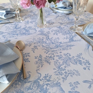 May include: A dining table set with a blue and white toile tablecloth featuring a floral and bird pattern. Clear glasses, light blue napkins, and gold-edged plates are arranged. A vase of pink roses is in the centre.