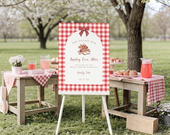 May include: A rectangular sign with a red and white gingham border and a white arched center. The text reads: SHE FOUND HER Appley Ever After WELCOME TO THE BRIDAL SHOWER HONORING Karly Tate 10.17.26. Above the text is a small illustration of an apple basket.