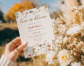 May include: A bridal shower invitation with the text "Love in Bloom" and "Bridal Shower". The invitation features floral illustrations in pink, yellow, and green. The invitation is held in front of a bouquet of white and beige flowers.