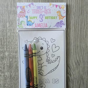 May include: A birthday coloring set featuring a dinosaur theme. The set includes a coloring page with a dinosaur illustration and three crayons. The packaging reads "She's a Three-Rex Happy 3rd Birthday Amelia."