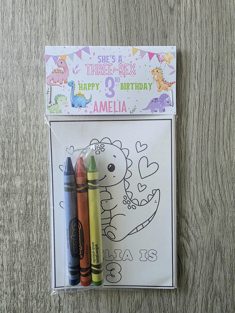May include: A birthday coloring set featuring a dinosaur theme. The set includes a coloring page with a dinosaur illustration and three crayons. The packaging reads "She's a Three-Rex Happy 3rd Birthday Amelia."