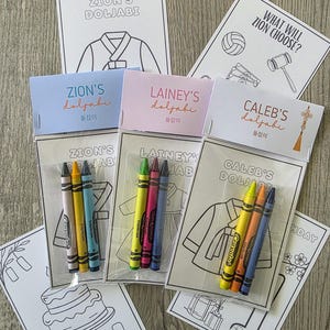 May include: Three sets of personalized Doljabi activity kits, each containing three Crayola crayons and a coloring sheet featuring a hanbok. The headers are labeled Zion's, Lainey's, and Caleb's Doljabi, with additional coloring pages showing a cake, a number one, and various symbols like a gavel and volleyball.