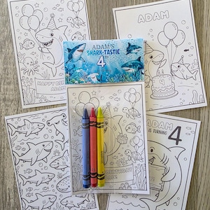 May include: A set of five white paper coloring sheets featuring shark-themed birthday illustrations with the text Adam's Shark-Tastic 4th Birthday, accompanied by a clear plastic pouch containing three crayons in blue, red, and yellow colors.