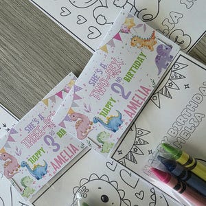 May include: A collection of children's birthday party favors featuring dinosaur-themed coloring pages and crayon packs. The favors include personalized labels with the name "Amelia" and the phrases "Happy Birthday" and "She's a Two-Rex" or "She's a Three-Rex".