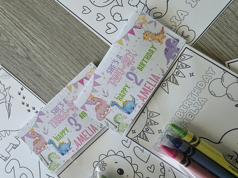 May include: A collection of children's birthday party favors featuring dinosaur-themed coloring pages and crayon packs. The favors include personalized labels with the name "Amelia" and the phrases "Happy Birthday" and "She's a Two-Rex" or "She's a Three-Rex".