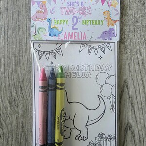 May include: A birthday coloring set featuring a coloring page with a dinosaur design and the text "HAPPY BIRTHDAY AMELIA". Includes three crayons in pink, blue, and green. The packaging reads "SHE'S A TWO-REX" and "HAPPY 2ND BIRTHDAY AMELIA".