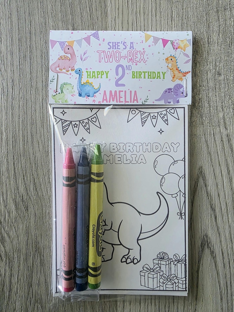 May include: A birthday coloring set featuring a coloring page with a dinosaur design and the text "HAPPY BIRTHDAY AMELIA". Includes three crayons in pink, blue, and green. The packaging reads "SHE'S A TWO-REX" and "HAPPY 2ND BIRTHDAY AMELIA".