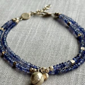 May include: A double-strand bracelet featuring faceted blue beads, small metallic cube spacers, and a round, textured metallic charm. The bracelet includes a metallic chain extender with a circular clasp component, showcasing a detailed, weathered finish.