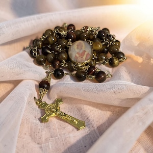 May include: A rosary featuring dark brown and golden-brown tiger eye beads connected by gold-toned wire. The piece includes a circular pendant with a religious portrait and a gold-toned crucifix with intricate detailing at the base of the beads.