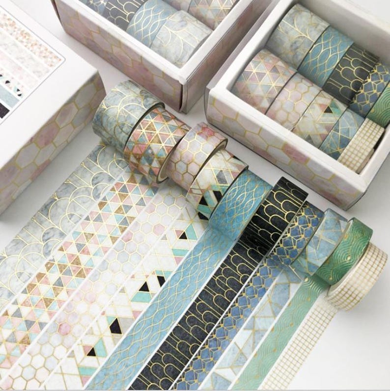 Pu&ograve; includere: Un set di 10 nastri washi decorativi con motivi geometrici in tonalit&agrave; di blu, verde e oro. I nastri sono disposti a ventaglio e sono esposti in una scatola bianca con un motivo a nido d'ape.