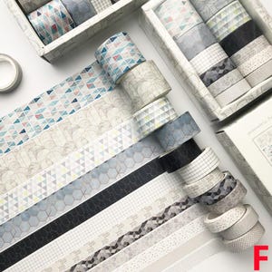 K&ouml;nnte beinhalten: Ein Set mit 12 Washi-B&auml;ndern mit geometrischen Mustern in Graut&ouml;nen, Wei&szlig; und Schwarz. Die B&auml;nder sind in einer Reihe angeordnet, wobei die Enden jedes Bandes sichtbar sind. Die B&auml;nder haben alle unterschiedliche Muster, aber sie alle haben eine &auml;hnliche minimalistische &Auml;sthetik.