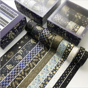 Pu&ograve; includere: Un set di 10 nastri washi con disegni dorati e blu su sfondo nero. I nastri sono disposti in una scatola con un motivo floreale. I nastri presentano disegni come stelle, pianeti, foglie e motivi geometrici.