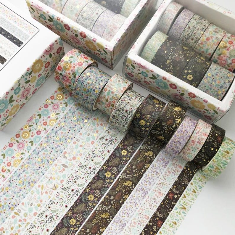 Pu&ograve; includere: Assortimento di nastri washi decorativi con motivi floreali e fogliari in tonalit&agrave; rosa, verde, marrone e oro. I nastri sono disposti in file su una superficie bianca.