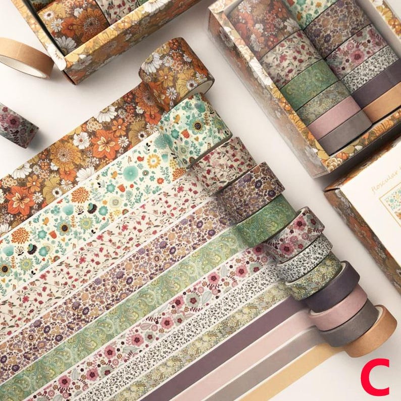 K&ouml;nnte beinhalten: Ein Set mit 12 floral gemusterten Washi-B&auml;ndern in verschiedenen Farben und Designs. Die B&auml;nder sind f&auml;cherf&ouml;rmig angeordnet und auf einer wei&szlig;en Oberfl&auml;che pr&auml;sentiert. Die B&auml;nder sind alle 15 mm breit und aus hochwertigem Papier gefertigt.