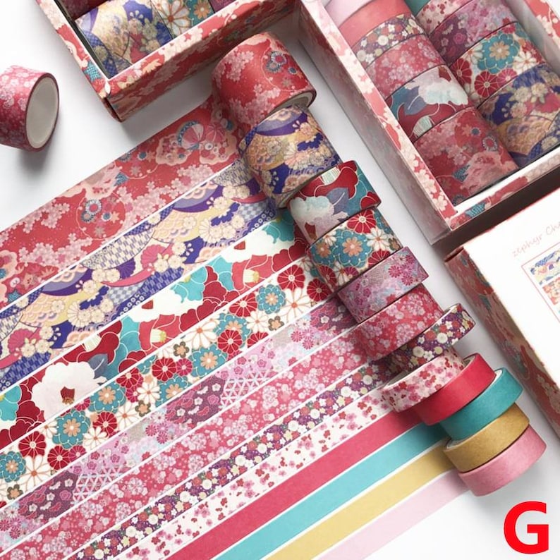 K&ouml;nnte beinhalten: Ein Set mit 10 Washi-B&auml;ndern mit floralen und geometrischen Mustern in Rot-, Rosa-, Blau- und Gr&uuml;nt&ouml;nen. Die B&auml;nder sind in einer Reihe auf einer wei&szlig;en Oberfl&auml;che angeordnet. Die B&auml;nder sind in einer Schachtel verpackt, auf der "Washi Tape" steht.