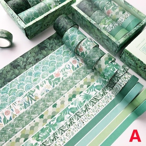 K&ouml;nnte beinhalten: Sortiment gr&uuml;ner Washi-B&auml;nder mit verschiedenen floralen und geometrischen Mustern. Die B&auml;nder sind f&auml;cherf&ouml;rmig angeordnet und in zwei Schachteln mit den Aufschriften "Green Forest" und "Glitter Forest" pr&auml;sentiert.