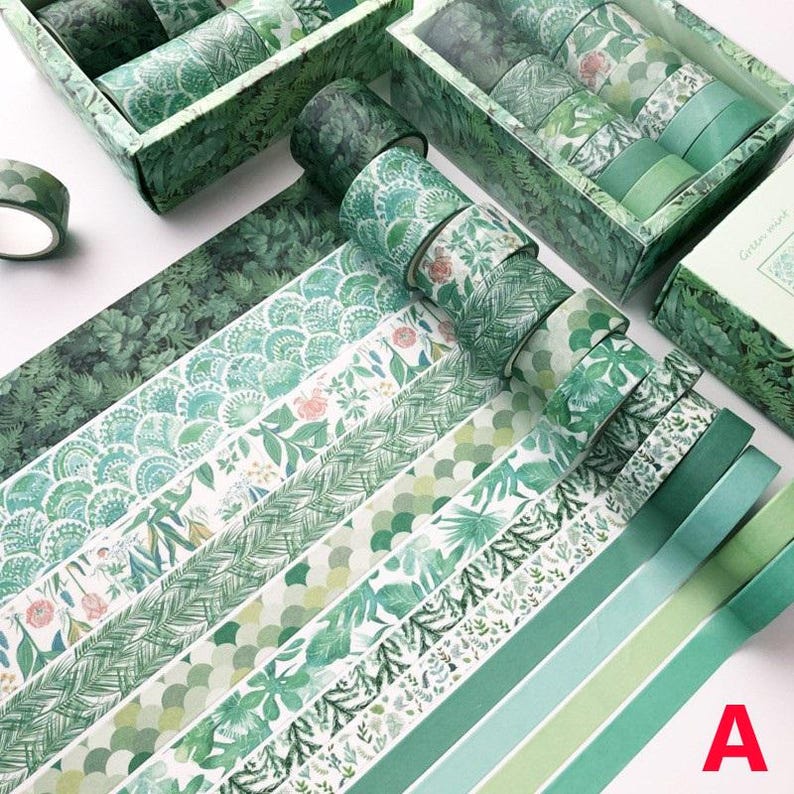 K&ouml;nnte beinhalten: Sortiment gr&uuml;ner Washi-B&auml;nder mit verschiedenen floralen und geometrischen Mustern. Die B&auml;nder sind f&auml;cherf&ouml;rmig angeordnet und in zwei Schachteln mit den Aufschriften "Green Forest" und "Glitter Forest" pr&auml;sentiert.