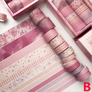 K&ouml;nnte beinhalten: Ein Set mit 10 rosa und wei&szlig;en gemusterten Washi-B&auml;ndern in einer rosa Box. Die B&auml;nder zeigen verschiedene Designs, darunter florale, geometrische und karierte Muster. Die Box ist mit "Cherry Flower" beschriftet.