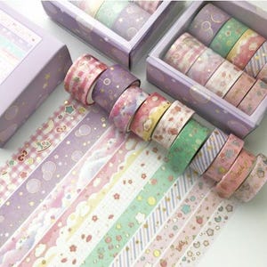 Pu&ograve; includere: Un set di 10 nastri washi colorati con vari motivi, tra cui stelle, nuvole e fiori. I nastri sono decorati con glitter dorati e sono confezionati in una scatola di plastica trasparente con un coperchio viola.