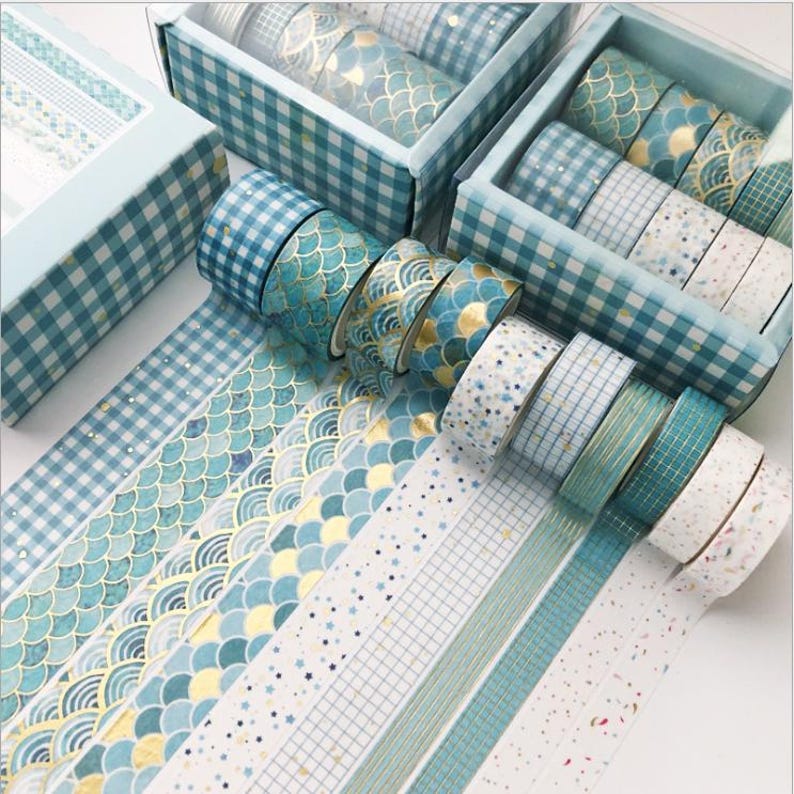 Pu&ograve; includere: Un set di nastri washi con vari motivi, tra cui squame blu e dorate, onde blu e dorate, a scacchi blu e bianchi e bianco con glitter blu e dorato. I nastri sono disposti in fila su una superficie bianca.