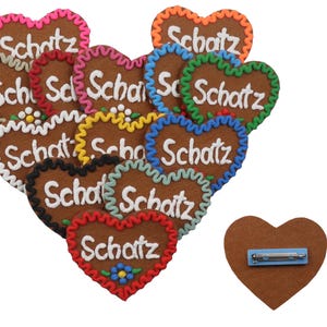 May include: A collection of heart-shaped gingerbread cookies with the word "Schatz" in white icing. Each cookie has a different colored border, including pink, orange, green, and blue. One cookie has a pin on the back.